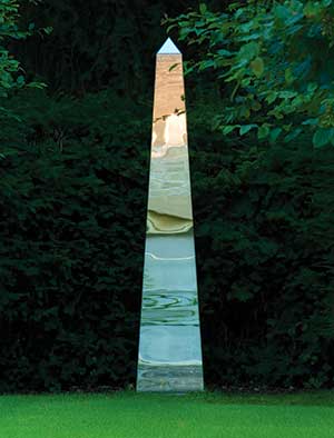 Gleaming stainless steel obelisk contrasting with dark planting behind